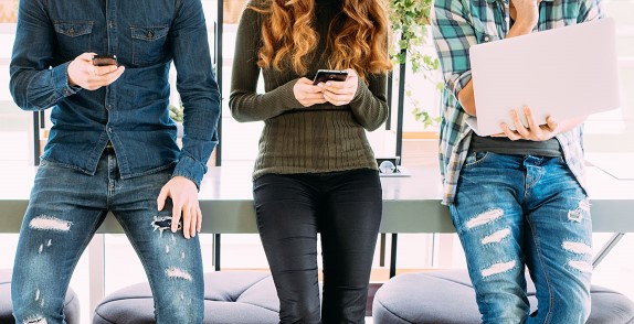 Three people sat in a row using phones and a laptop