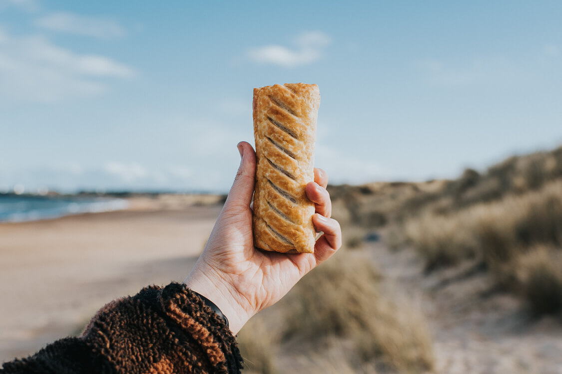 Hand holding Greggs sausage roll at the beach