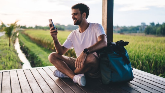 Man looking at his phone. He's sitting on a decking, with corn fields in the background